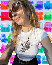 Load image into Gallery viewer, A woman wearing a white crewneck t-shirt with a black butterfly graphic on the front, styled with dark sunglasses and a silver necklace. She is posed in front of a repeating background of colorful, translucent plastic chair-like shapes.